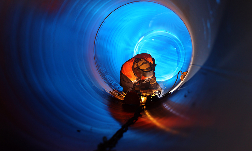 A fully covered and protected worker welds inside a large pipe, using an industrial flashlight to see